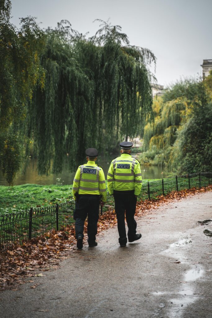 Two police officers in high-visibility jackets walking along a path in a lush park on a bright day.